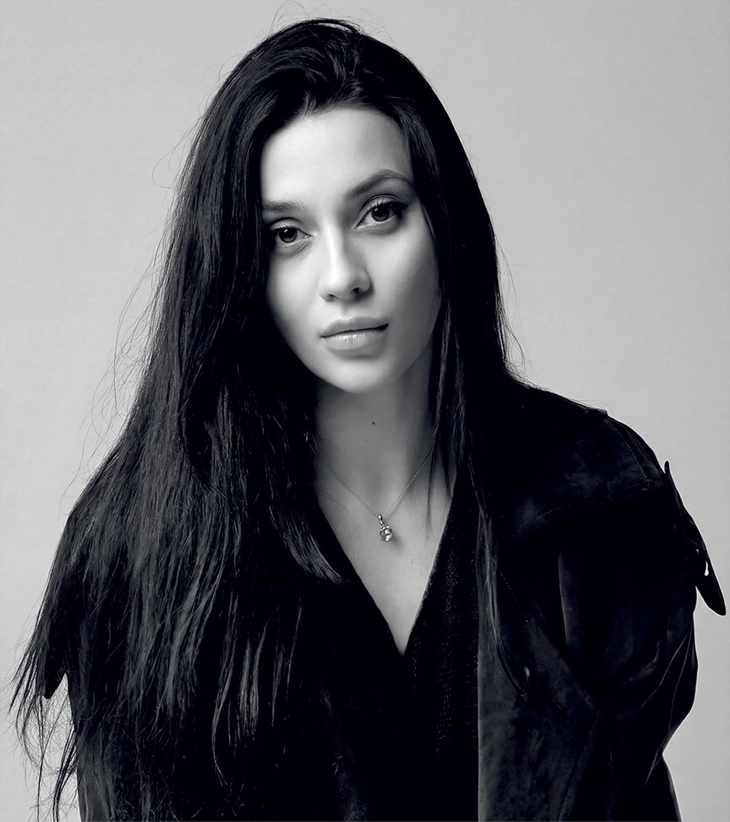 Woman with long dark hair and a neutral expression wearing a dark jacket and necklace, photographed against a plain light background.