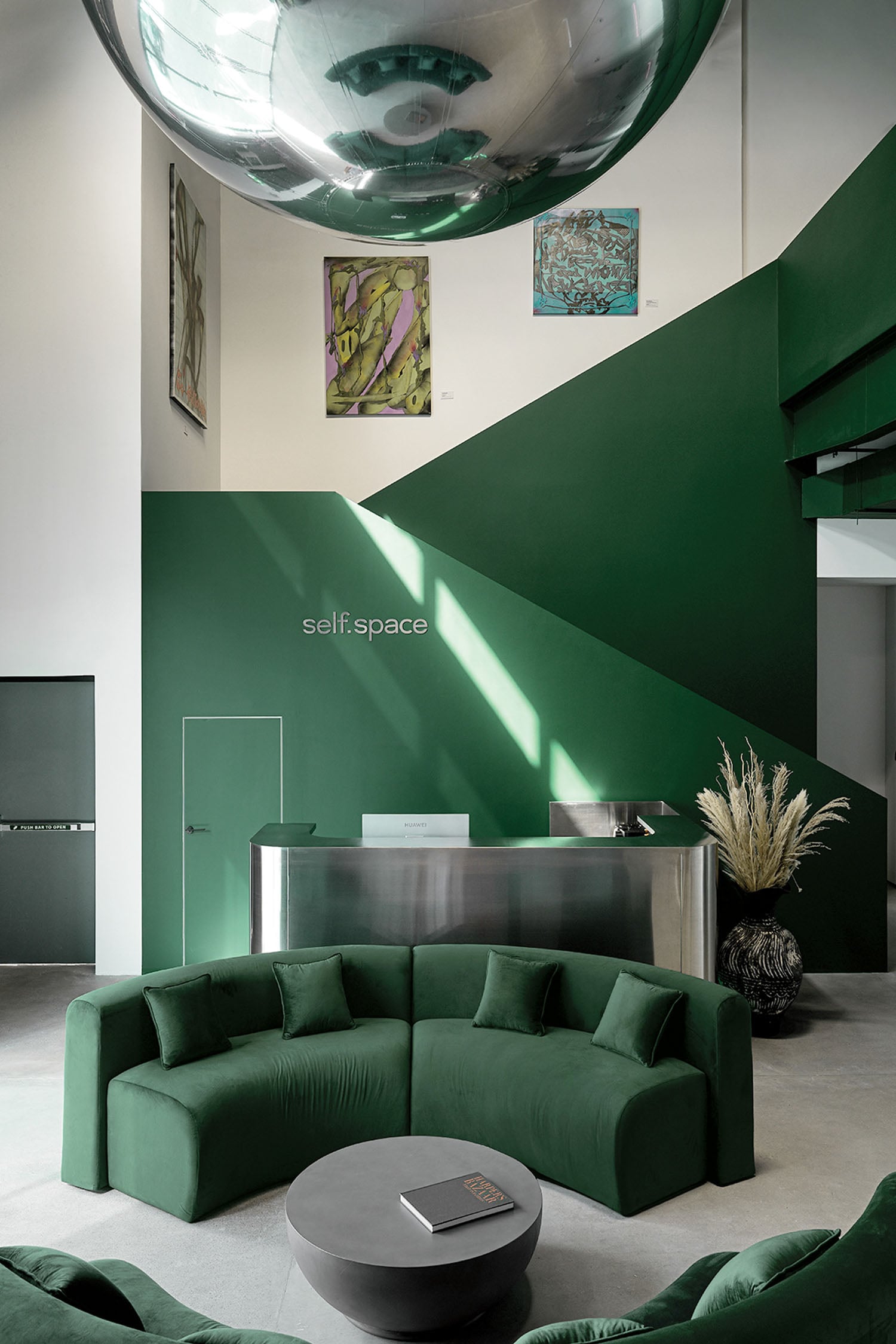 Modern lobby with green curved sofas, a stainless steel reception desk, green accent walls, abstract art, and a large reflective sphere overhead.