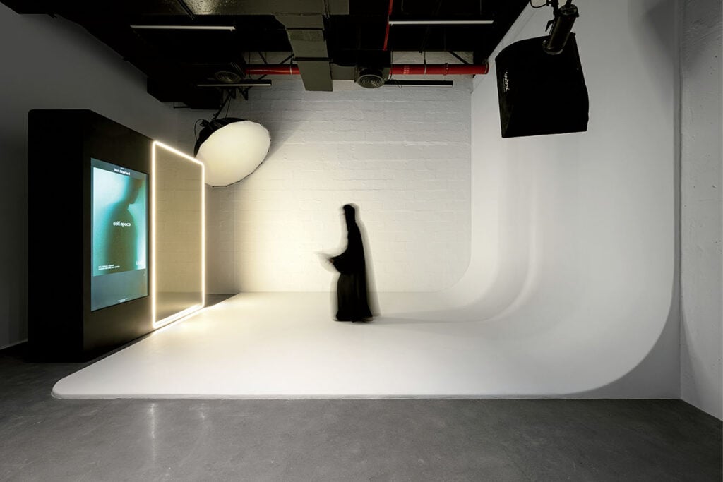 A person in a black abaya stands blurred in a minimalist photography studio with curved white walls, soft lighting, and a large illuminated screen.