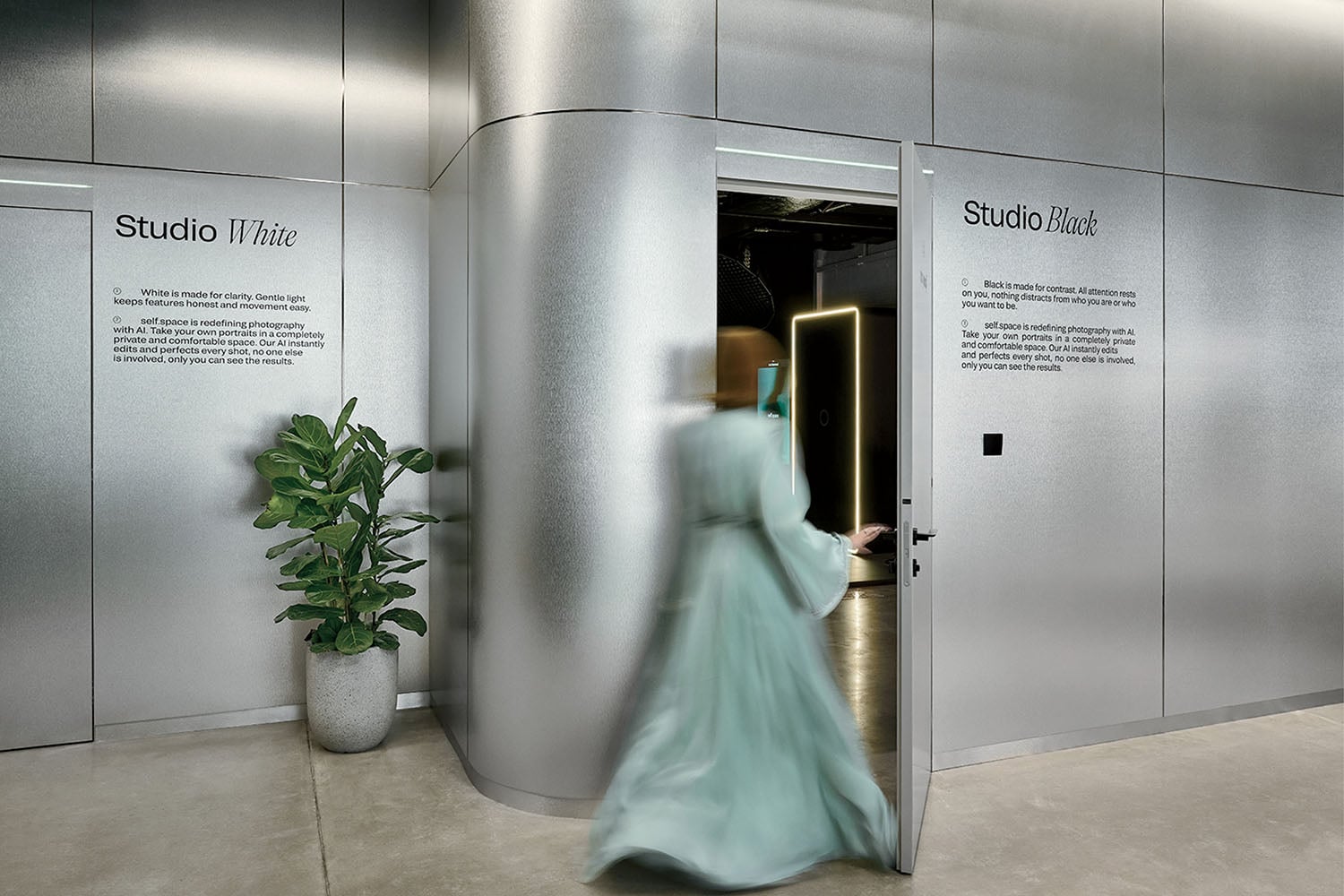 A person in a light blue dress enters a modern space with metallic walls, passing by a potted plant; signs read "Studio White" and "Studio Black.