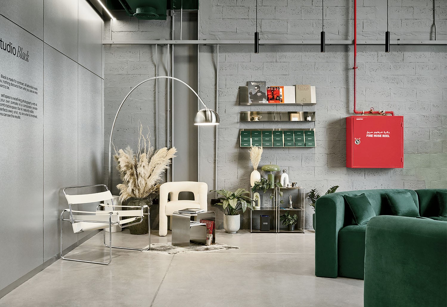 Modern lounge area with green sofa, white chairs, plants, shelves with books, and a red fire hose reel mounted on a gray concrete wall.