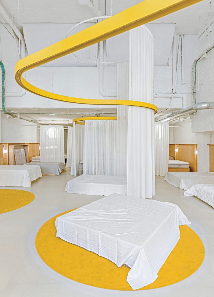 A modern, white room with several beds covered in white sheets, separated by white curtains hanging from circular yellow ceiling tracks.