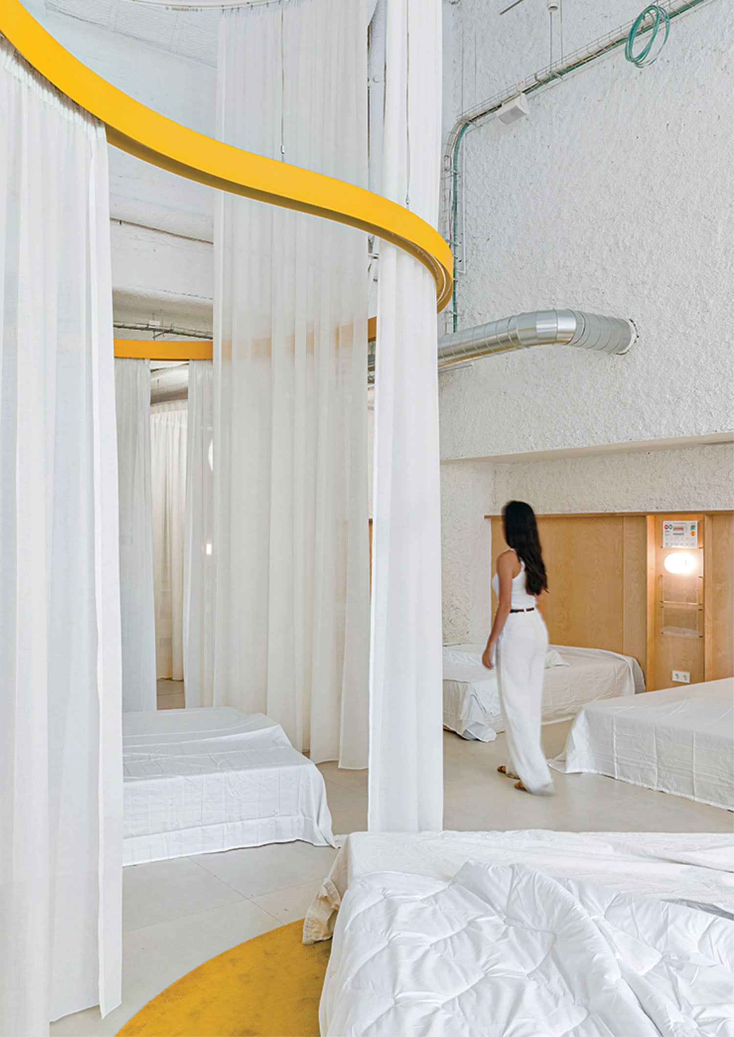 A woman in white walks through a minimalist room with white beds, sheer curtains, exposed ducts, and a yellow curved ceiling feature.