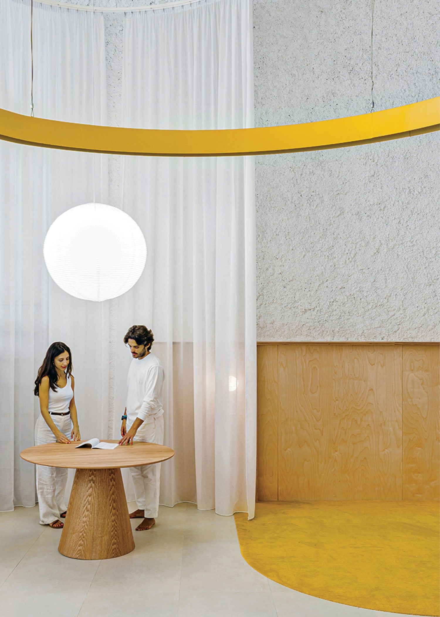 Two people stand at a round wooden table in a modern, minimalist room with white curtains, a textured wall, and a large round light fixture hanging above.