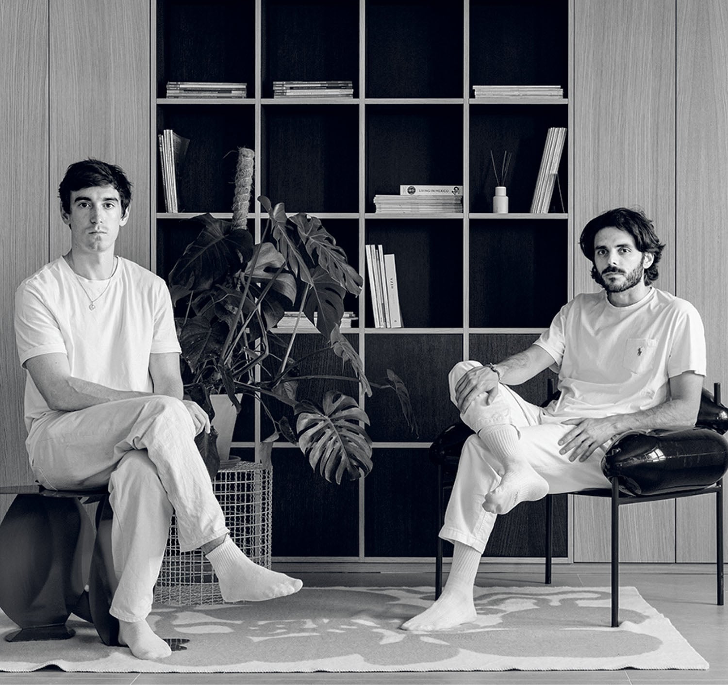 Two men in white clothing sit barefoot in a modern room with a large bookshelf, a plant, and minimal decor.
