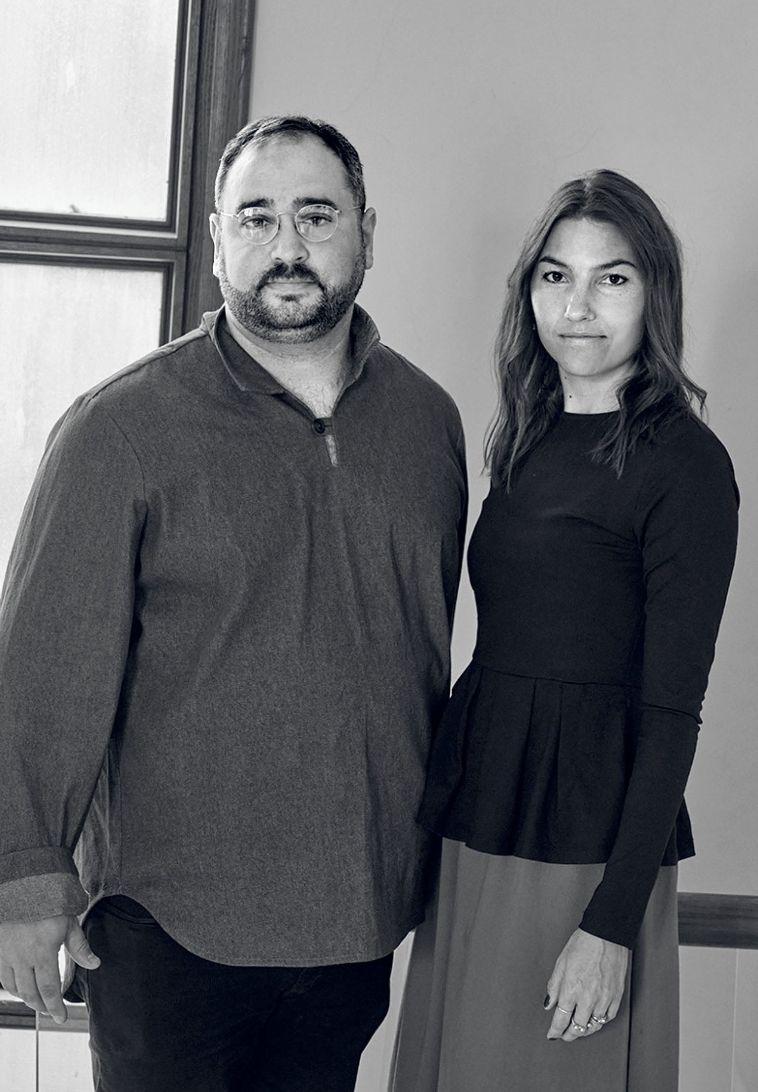 A man and a woman stand side by side indoors near a window, both looking at the camera with neutral expressions.