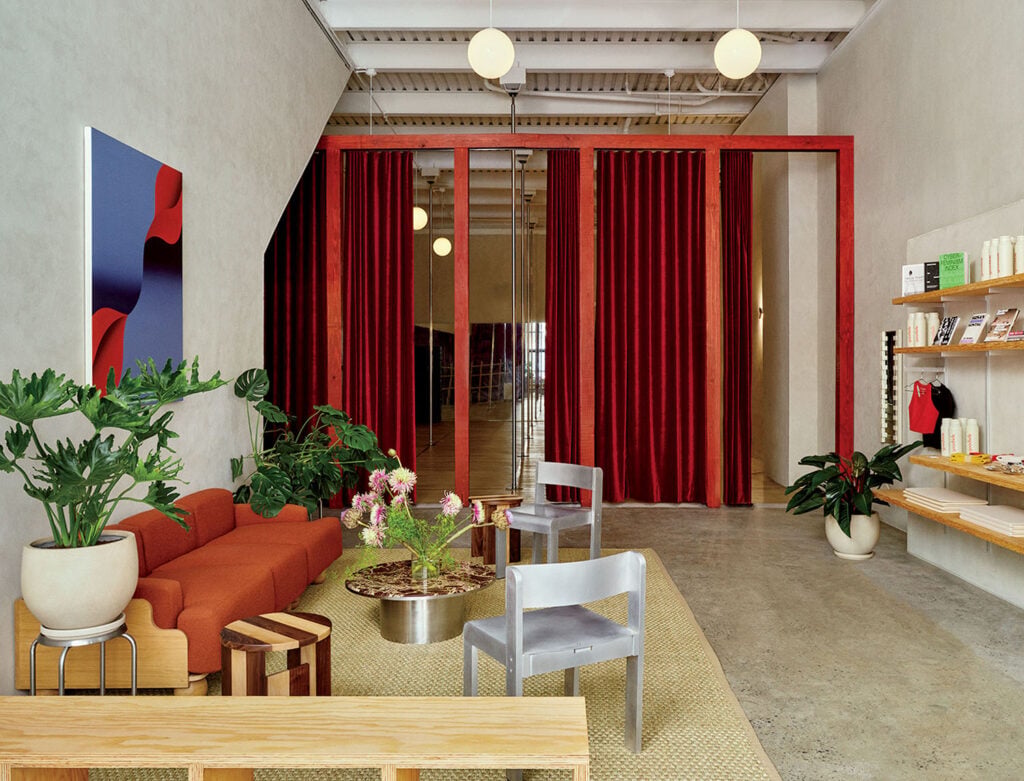 Modern waiting area with a red sofa, silver chairs, potted plants, a low table, wall art, red curtains, and shelves with books and products on a concrete floor.