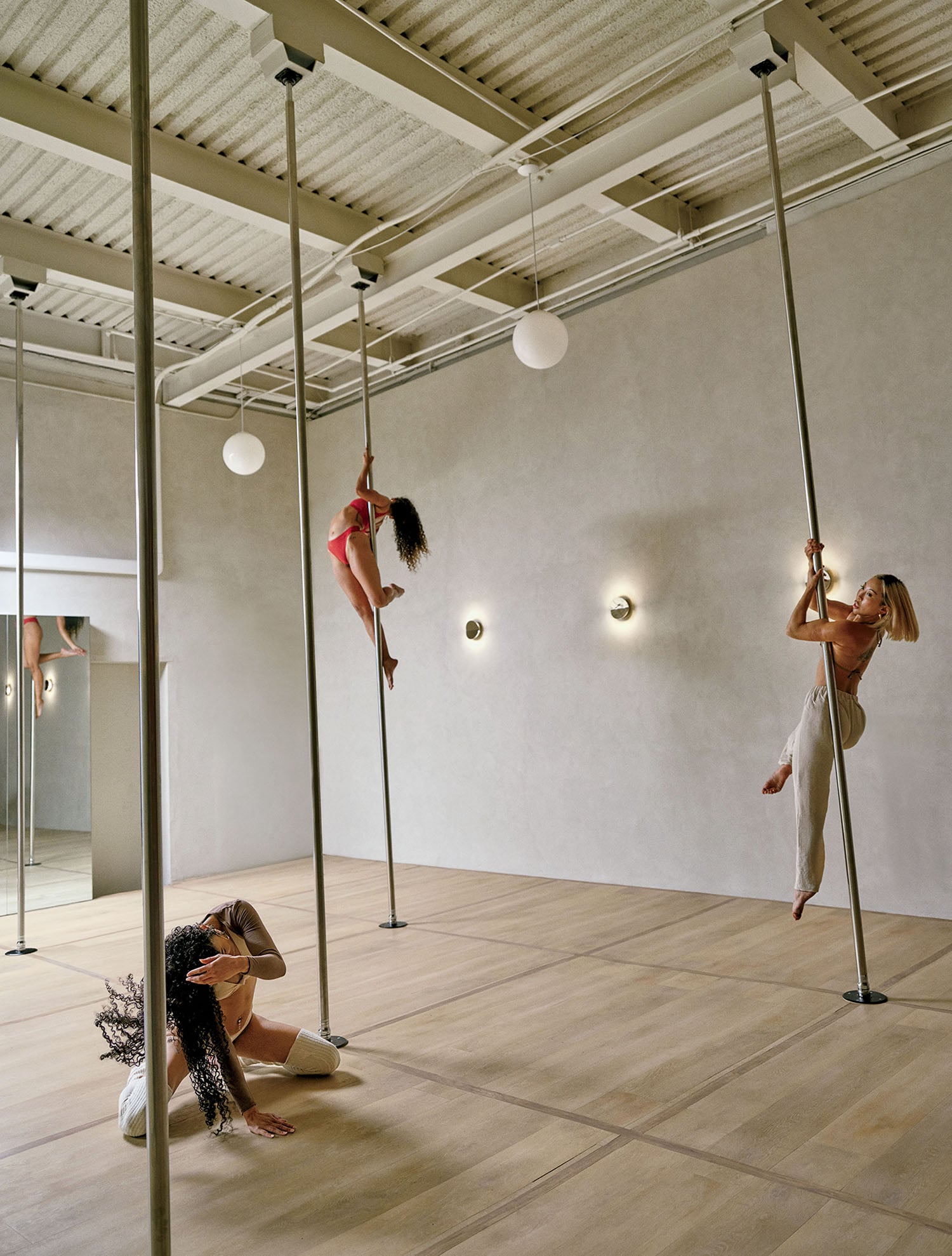 Three women practicing pole dancing in a spacious studio with wooden floors and neutral walls. One is climbing, one poses mid-air, and one is crouched on the floor.