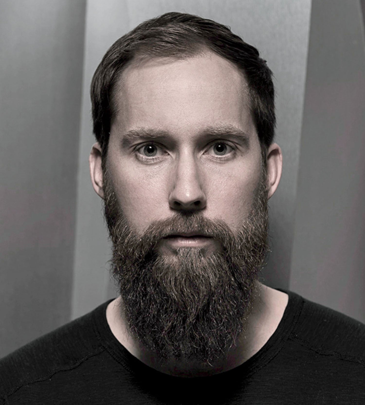 A man with a full beard and short hair, wearing a dark shirt, stands against a gray, geometric background and looks directly at the camera.
