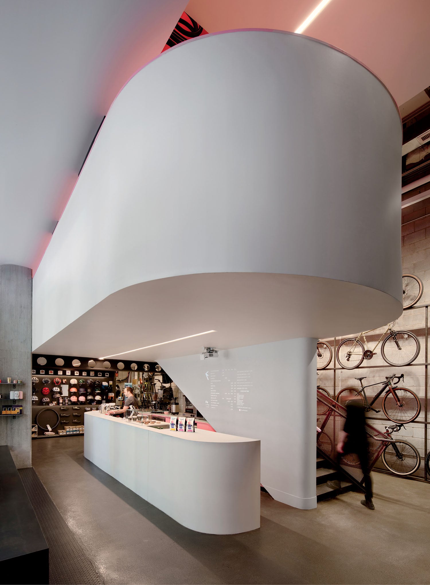 Modern bike shop interior with a curved white mezzanine, counter with products, wall-mounted bicycles, and a person ascending the stairs.
