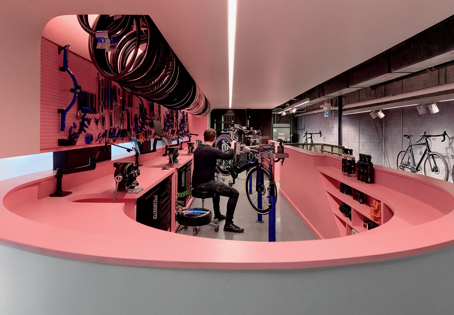 A person repairs a bicycle in a modern workshop with pink counters, wall-mounted tools, and bicycles hanging and displayed throughout the space.