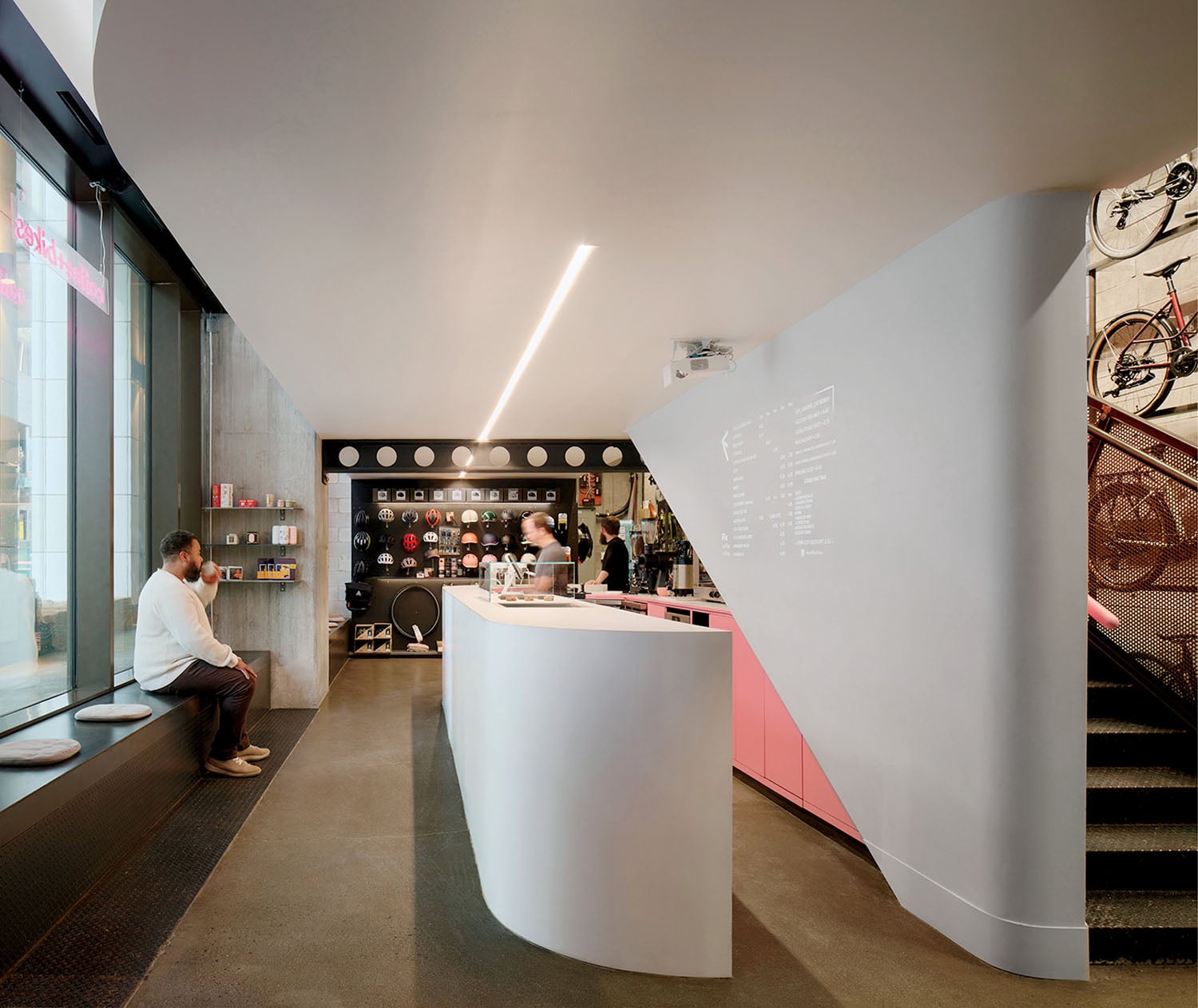 Modern café interior with curved white counter, menu projected on the wall, a seated person by the window, and bicycles mounted on the back wall.