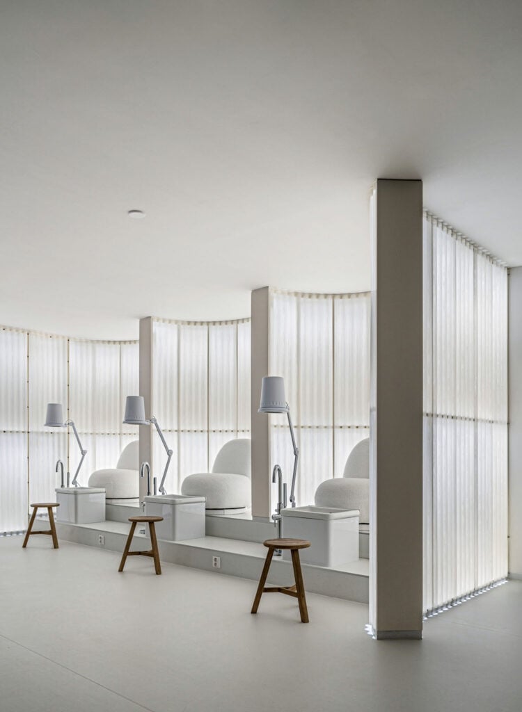 A minimalist pedicure spa room with three white chairs, individual lamps, wooden stools, and soft natural light filtering through sheer curtains.