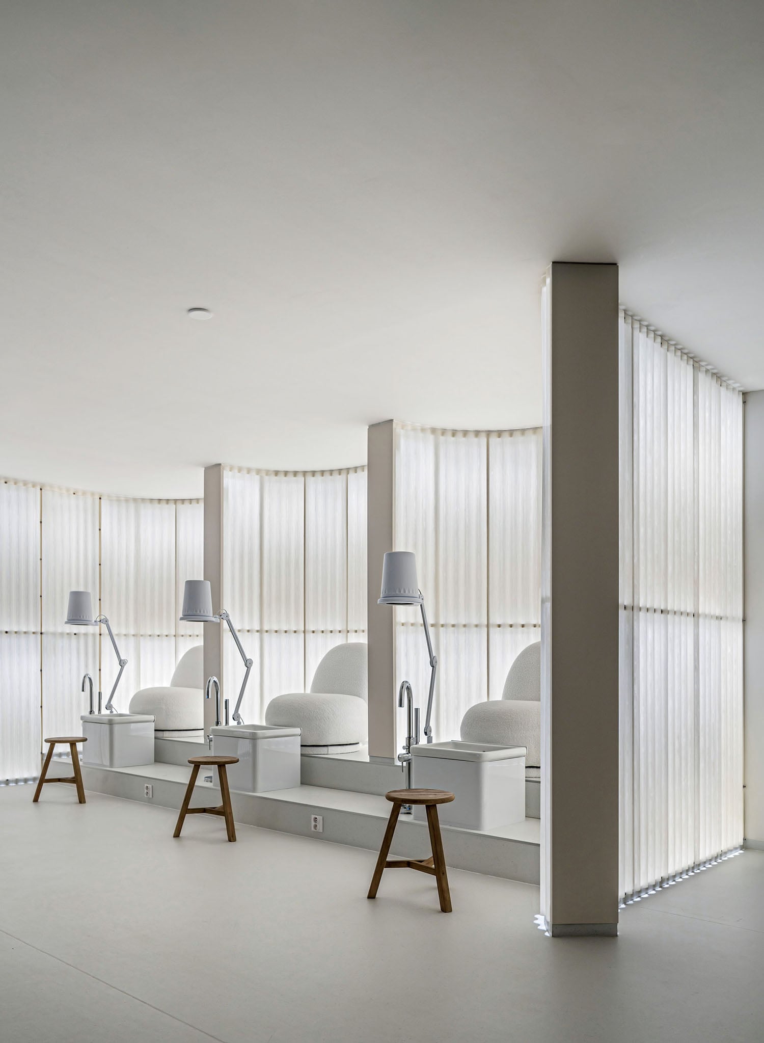 A minimalist pedicure spa room with three white chairs, individual lamps, wooden stools, and soft natural light filtering through sheer curtains.