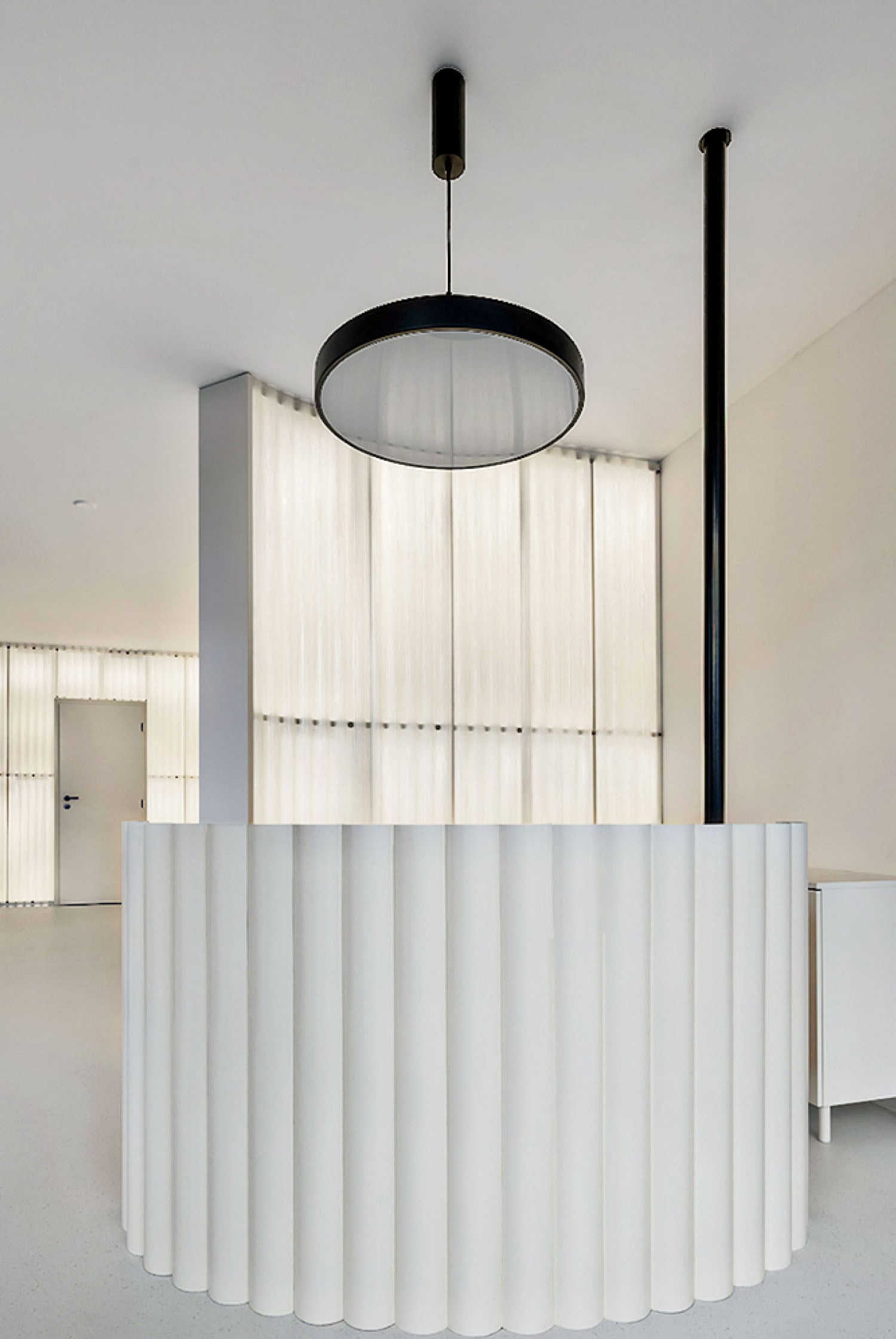 A modern reception desk with a curved, ribbed white facade stands beneath a large circular pendant light in a minimalist room with illuminated wall panels.