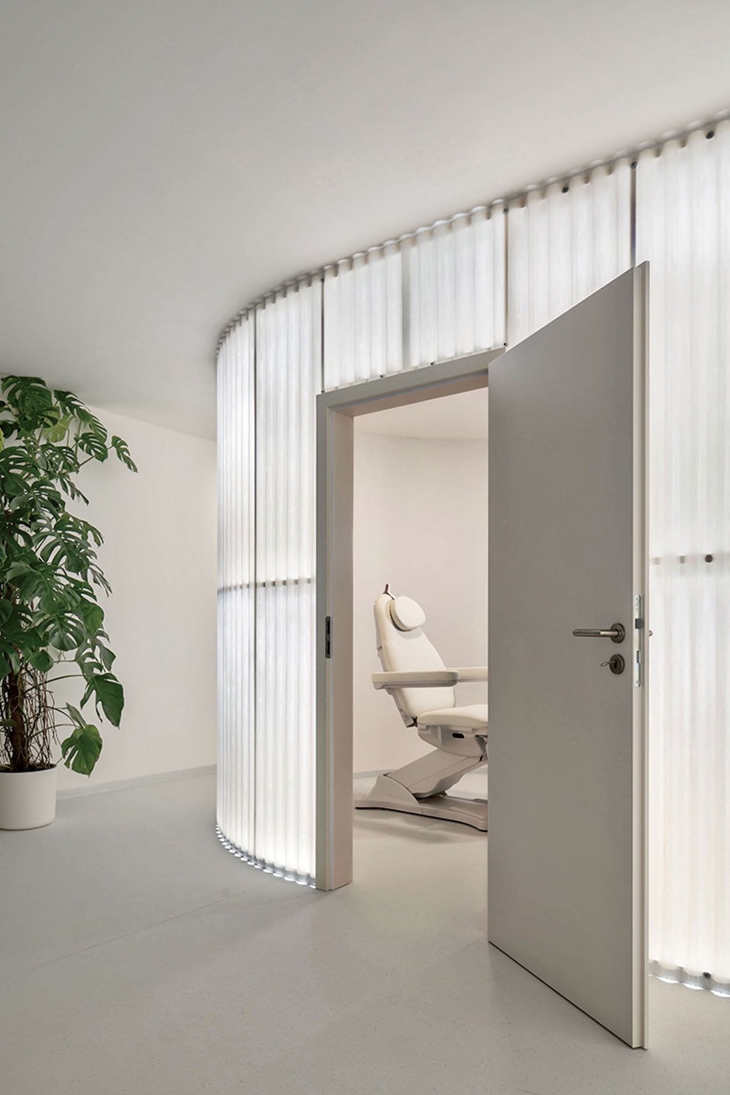 A modern, minimal clinic room with a white reclining chair is visible through a partially open door; illuminated vertical panels and a large potted plant are nearby.
