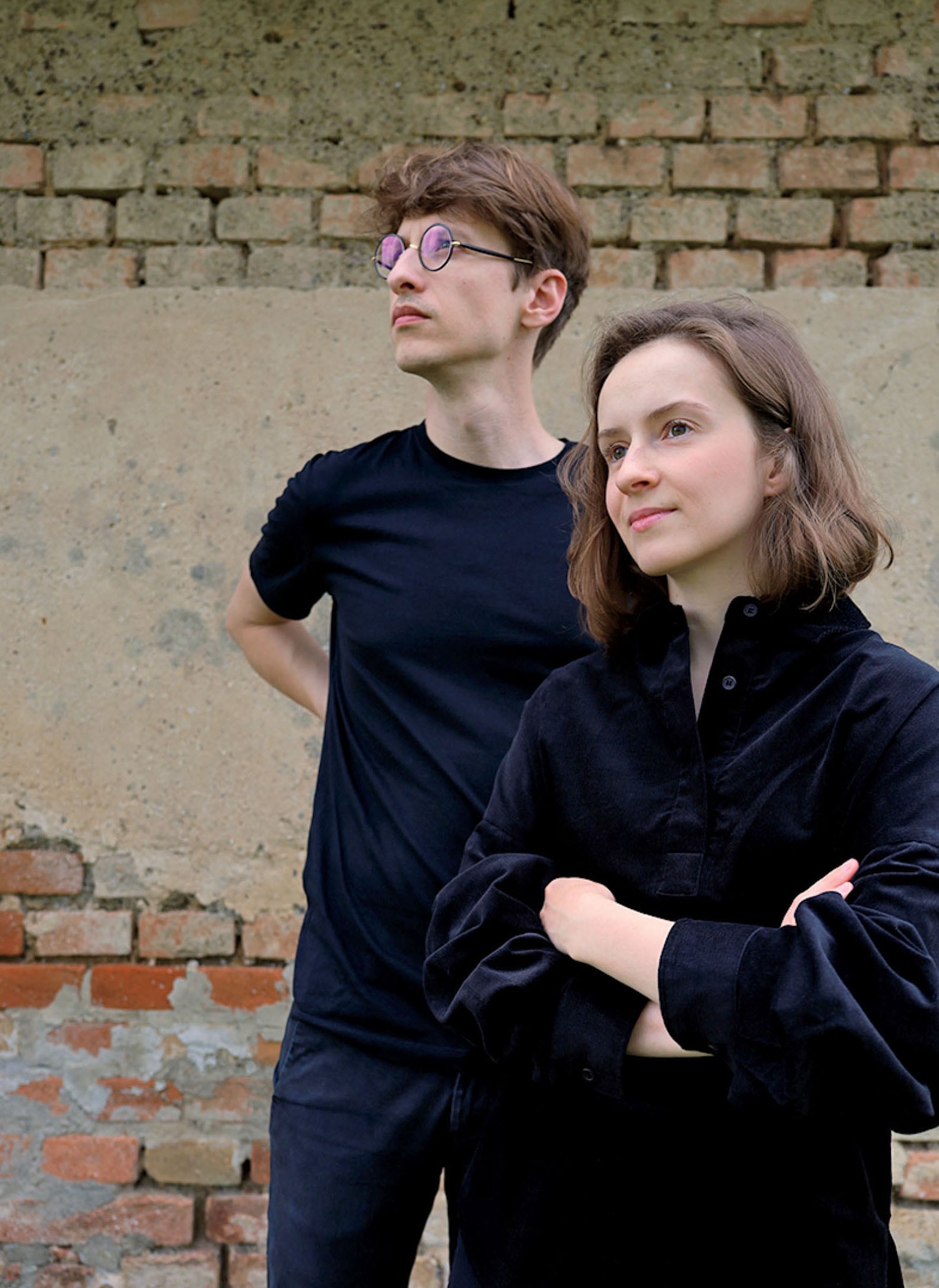 Two people stand in front of a brick and concrete wall, both wearing black tops. One has arms crossed and the other has hands behind their back, looking in different directions.