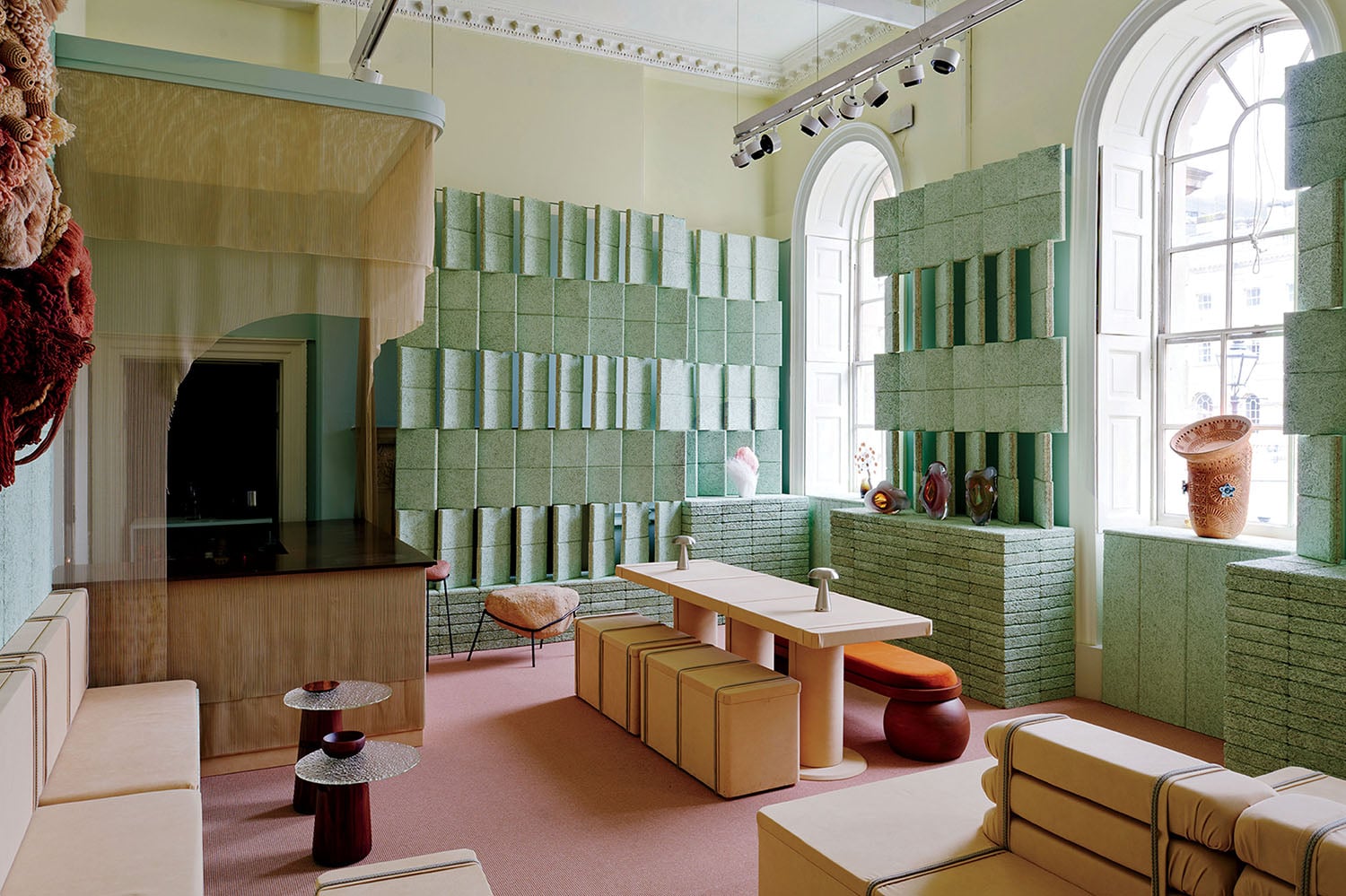 Modern room with pastel green textured walls, tall arched windows, beige modular seating, a central table, decorative vases, and soft pink carpeted flooring.