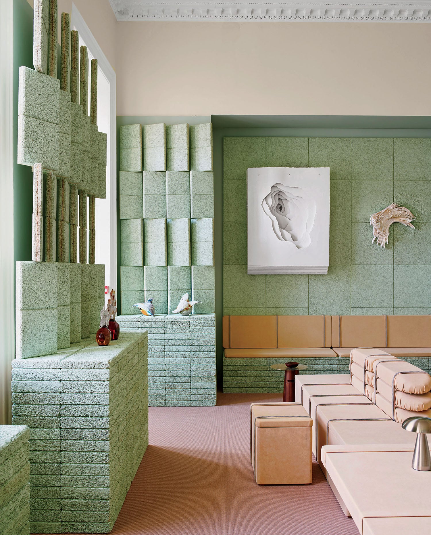 Modern room with green textured wall panels, beige modular seating, sculptures, and a framed artwork on the wall; the space features geometric and minimalist design elements.