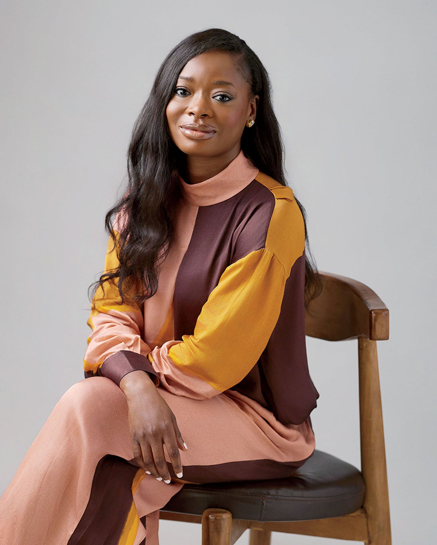 A woman with long, dark hair sits on a wooden chair, wearing a color-blocked, long-sleeved dress in shades of brown, pink, and yellow, against a plain light gray background.