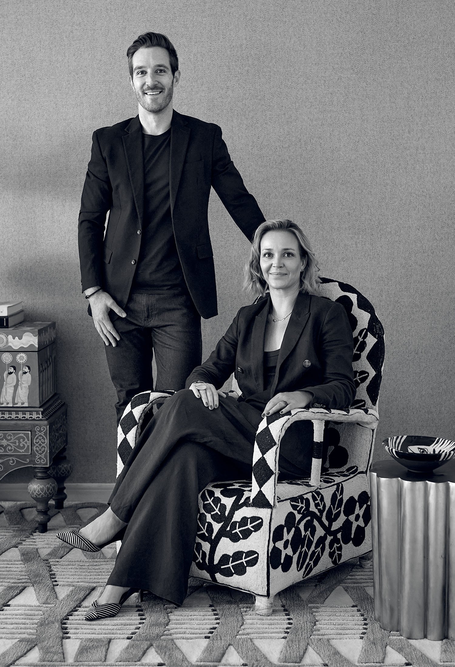 A man stands beside a seated woman in a patterned chair. Both wear dark clothing and pose in a modern, decorated room with patterned furniture and a rug.