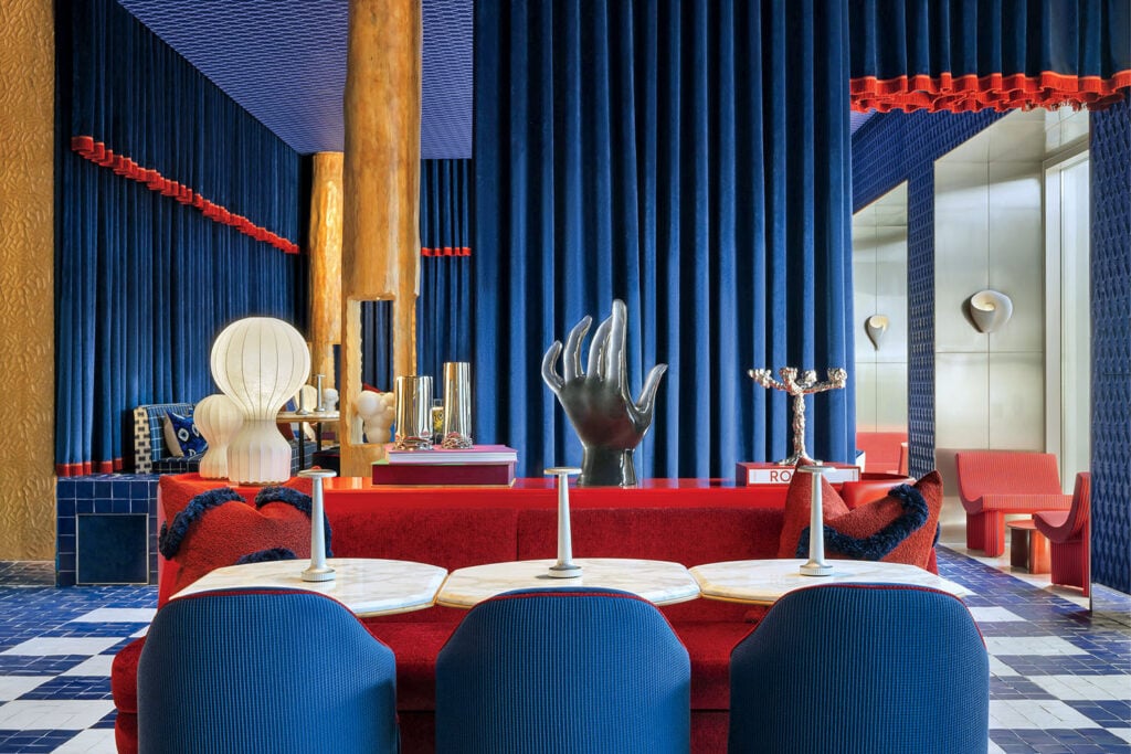 Bold, eclectic lounge with blue velvet drapes, red seating, marble tables, and assorted modern sculptures, including a large black hand and abstract lamp.