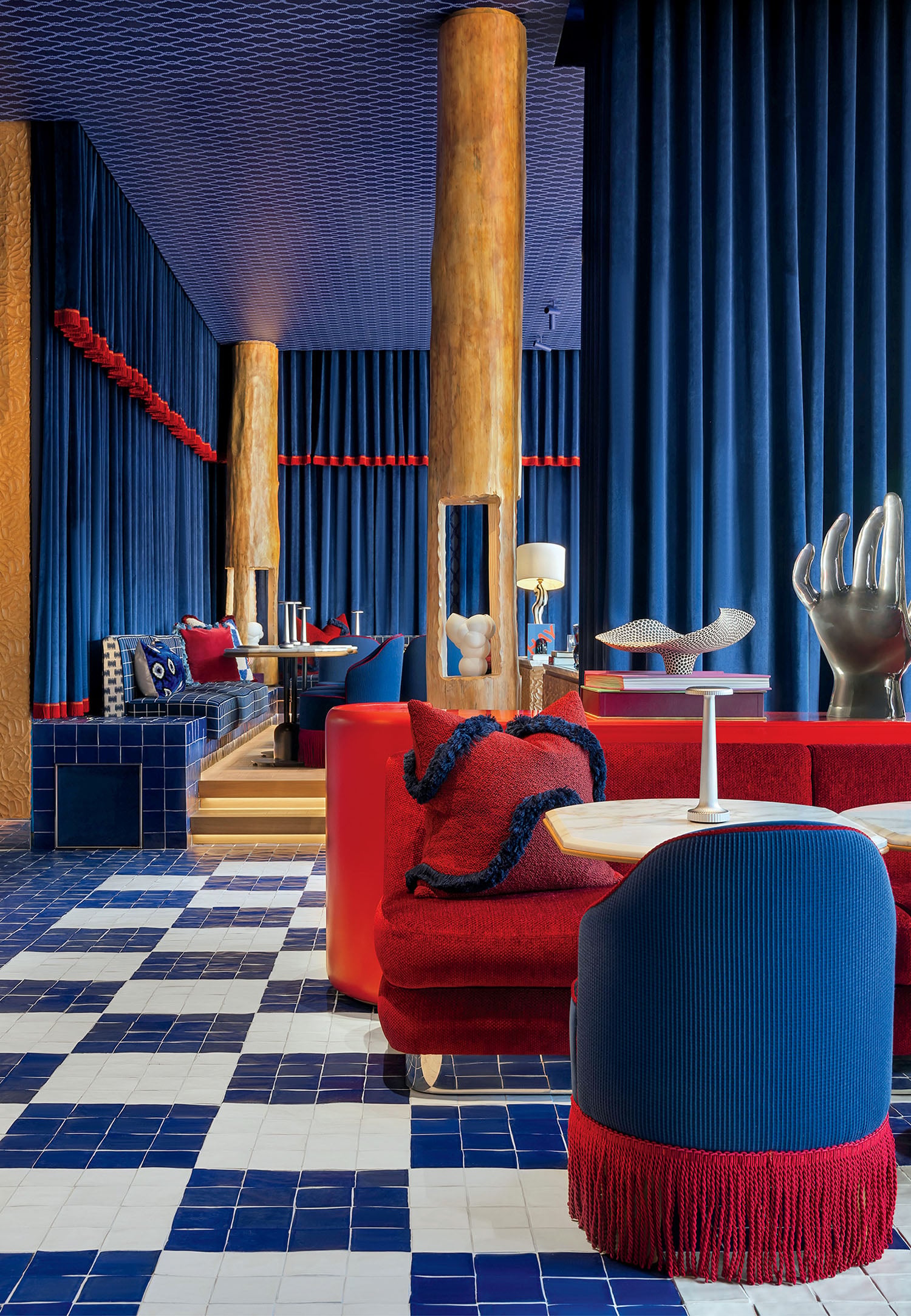 A modern lounge with blue and white checkered tile flooring, red and blue seating, wooden columns, and bold blue drapery.