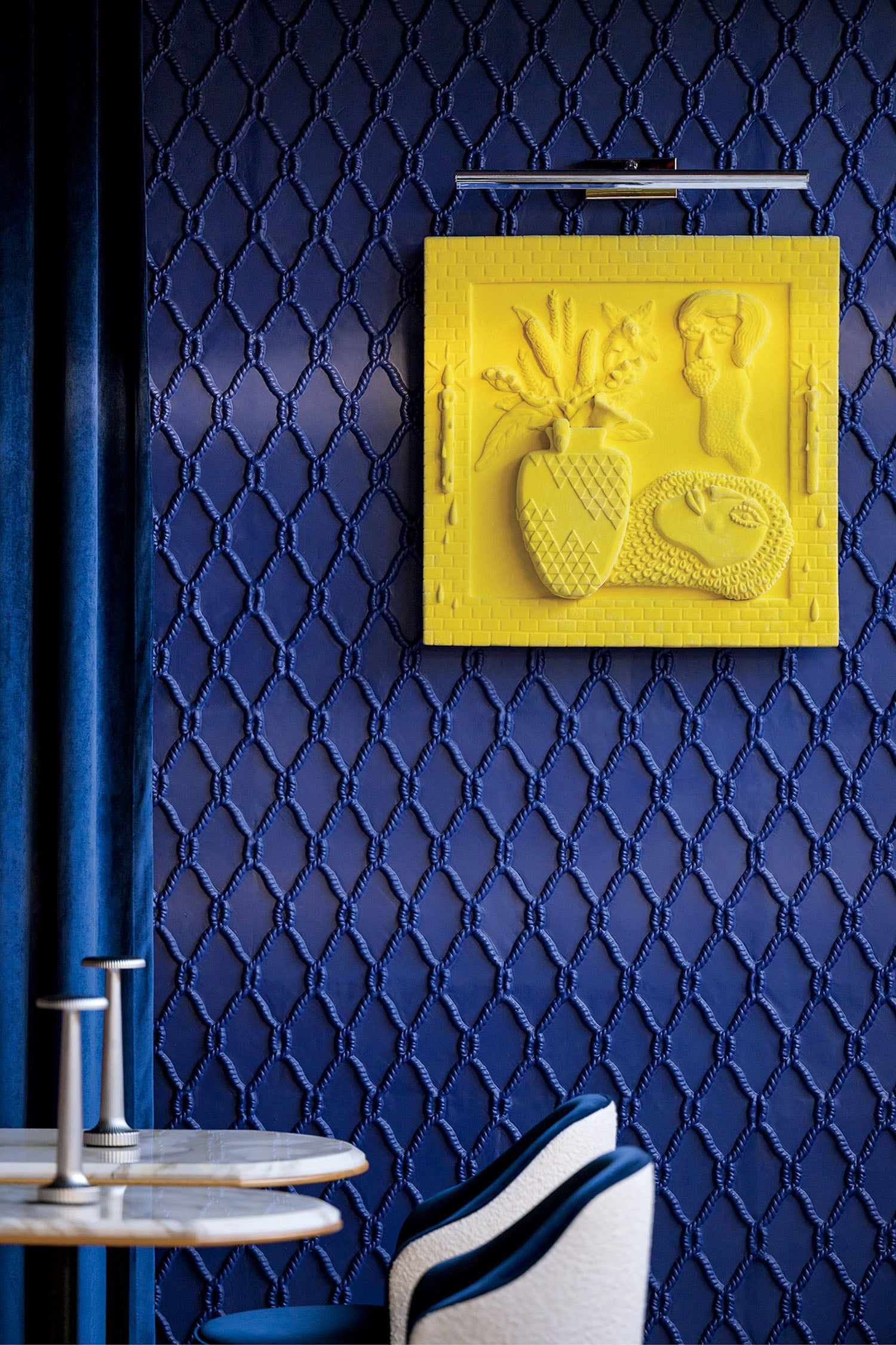 A blue wall with a patterned texture features a bright yellow relief artwork above a marble-topped table and blue upholstered chairs.