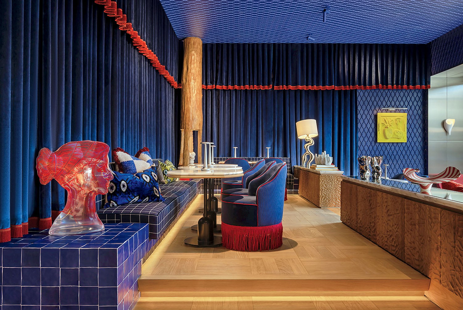 A stylish room with blue and red decor, velvet seating, wood accents, modern art pieces, and a mix of playful and sophisticated furnishings.