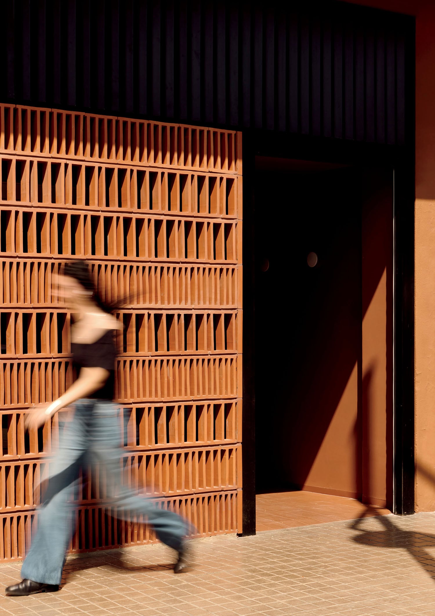 A person in motion walks past a patterned terracotta wall with sunlight casting shadows on the ground.