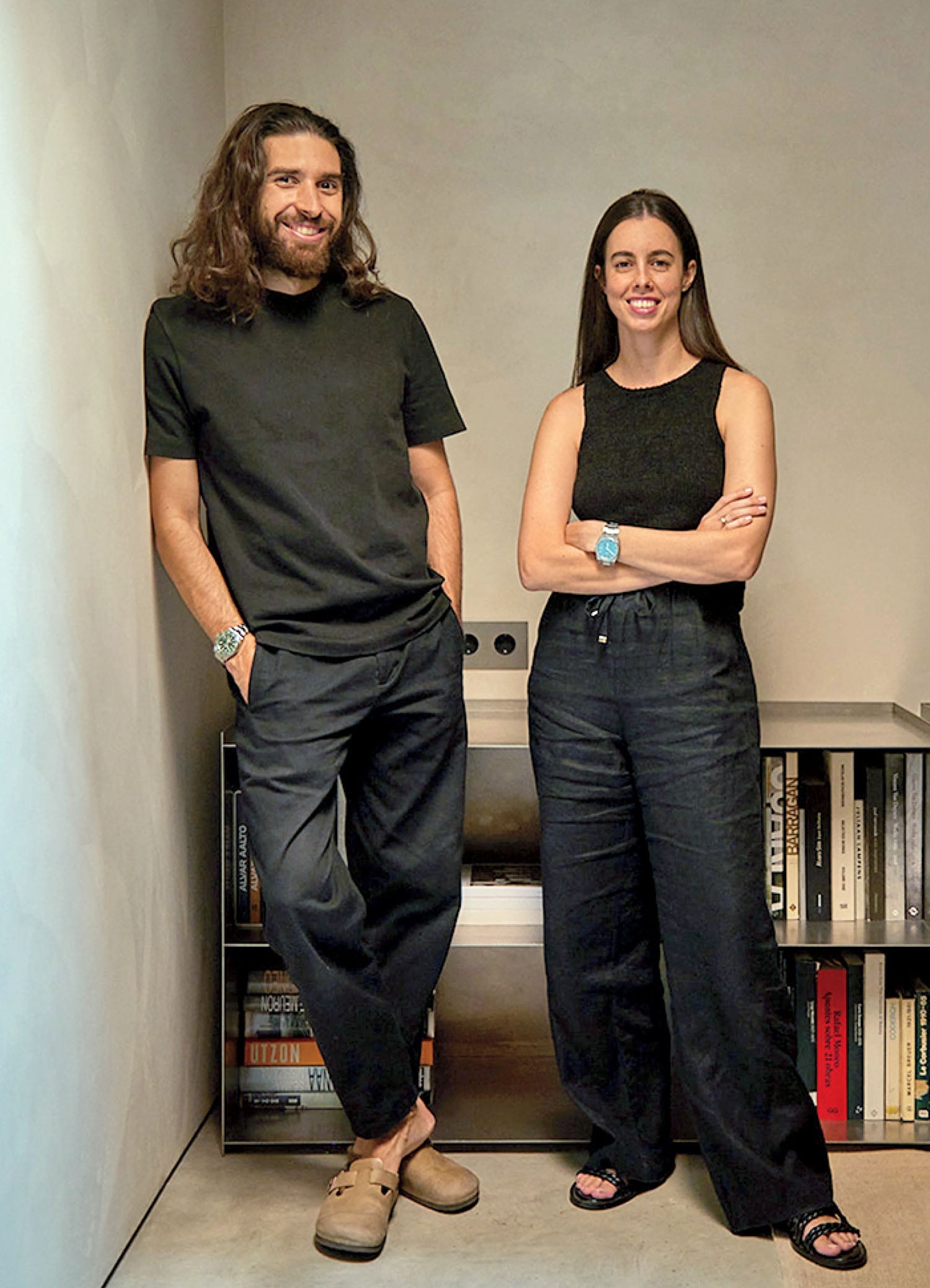 A man and woman stand side by side in a modern room, both dressed in black and smiling, with a bookshelf visible behind them.