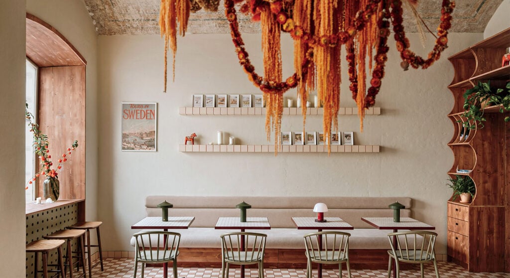 Cozy café interior with a long bench, tables, chairs, and patterned floor tiles. Dried floral arrangement hangs from the ceiling, with books and a Swedish travel poster on the walls.