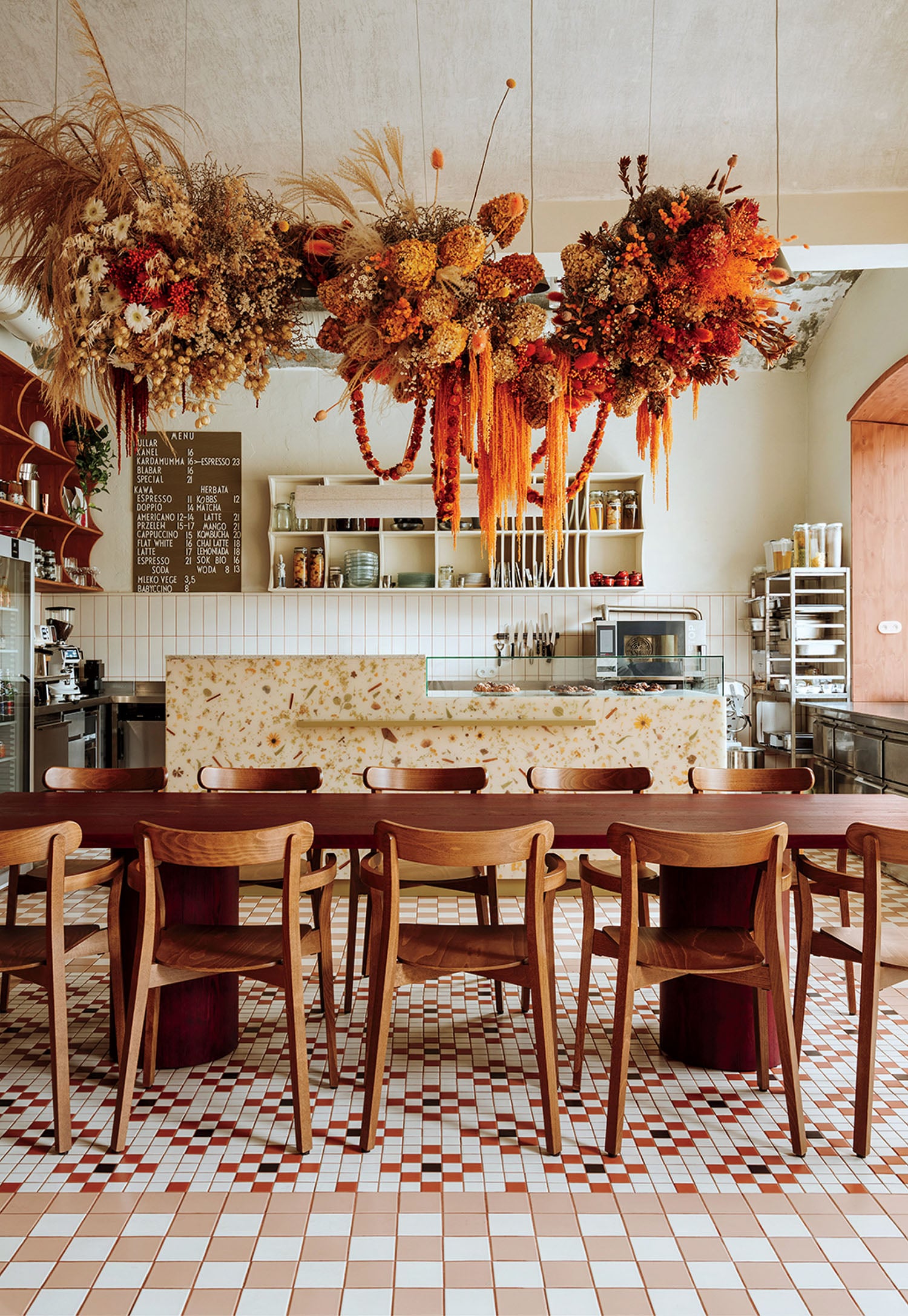 A modern café interior with wooden chairs, a tiled floor, a stone counter, and large floral arrangements hanging from the ceiling. Kitchen equipment and a menu board are visible in the background.