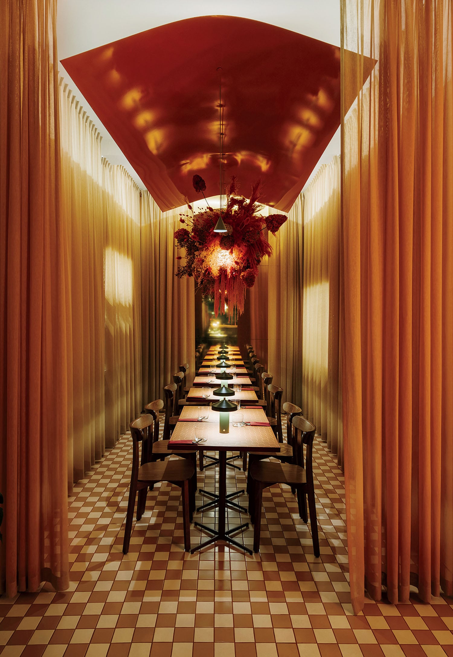 Long dining table with chairs in a narrow room, orange drapes, checkered tile floor, and a large central floral arrangement hanging from a red ceiling feature.