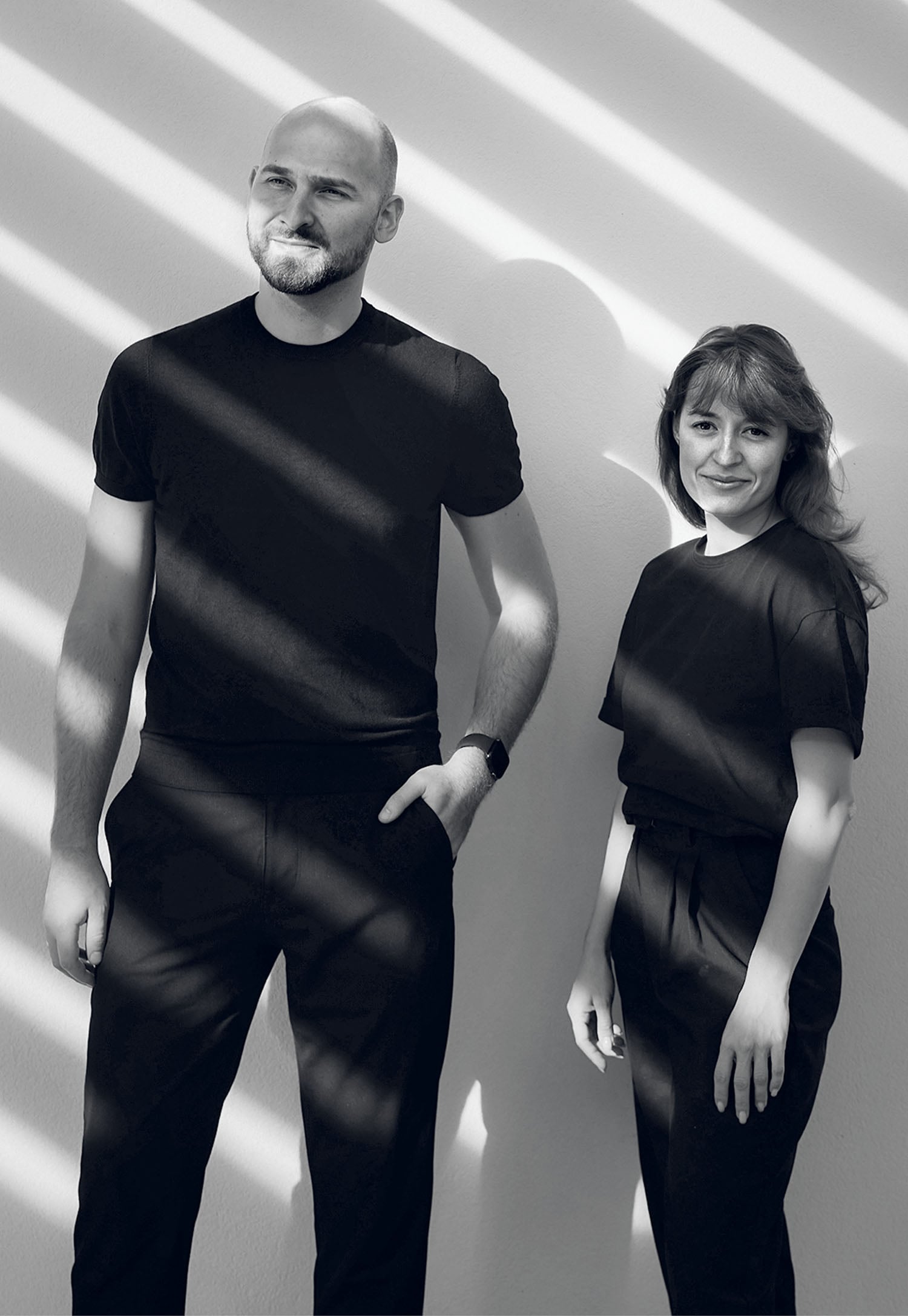 A man and a woman stand side by side against a light wall with diagonal shadows; both wear black outfits and look forward with neutral expressions.