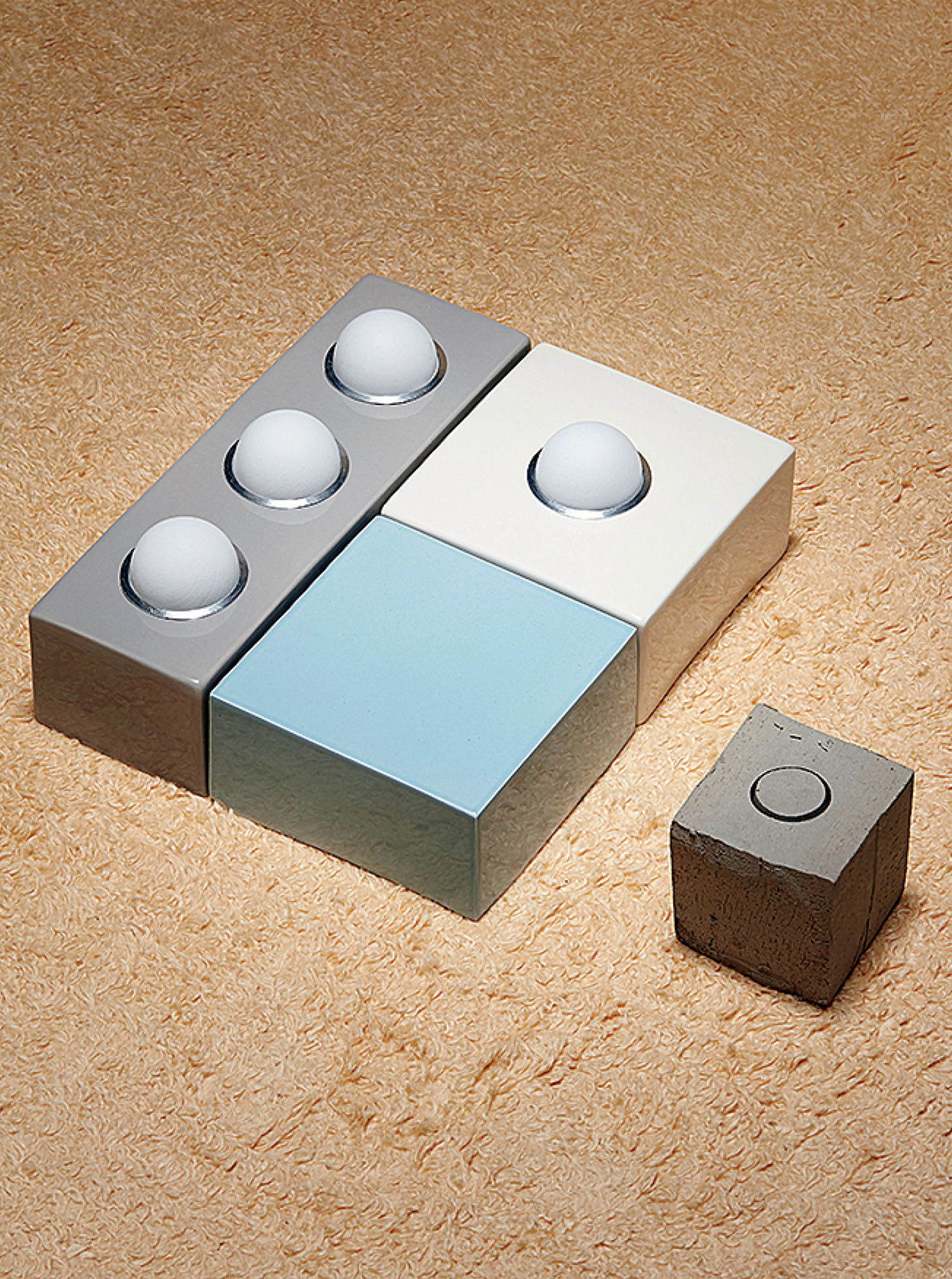 Four rectangular blocks, three with raised spheres and one plain, are arranged on a textured surface beside a separate small gray cube with a recessed circle on top.