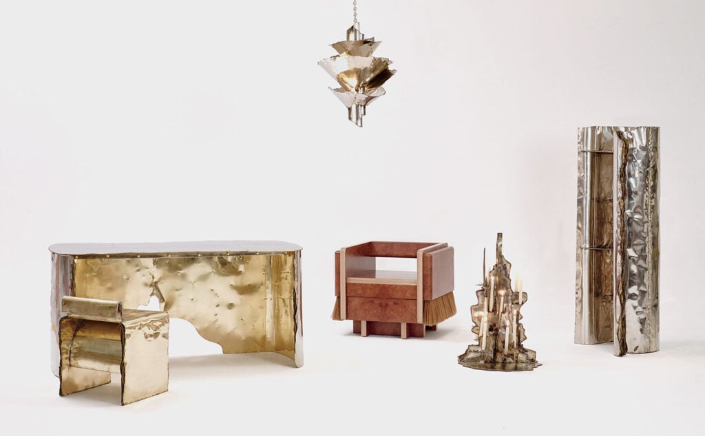 A set of modern, sculptural furniture pieces made of shiny metal and wood, including a desk, chair, chandelier, armchair, side table, and tall cabinet, arranged on a plain white background.