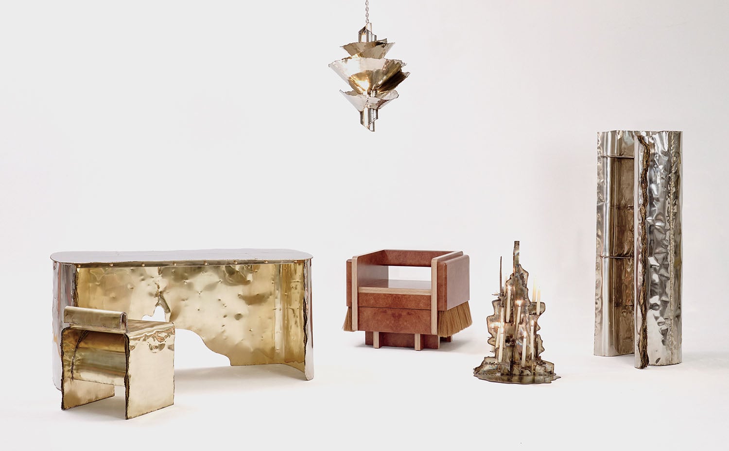 A set of modern, sculptural furniture pieces made of shiny metal and wood, including a desk, chair, chandelier, armchair, side table, and tall cabinet, arranged on a plain white background.