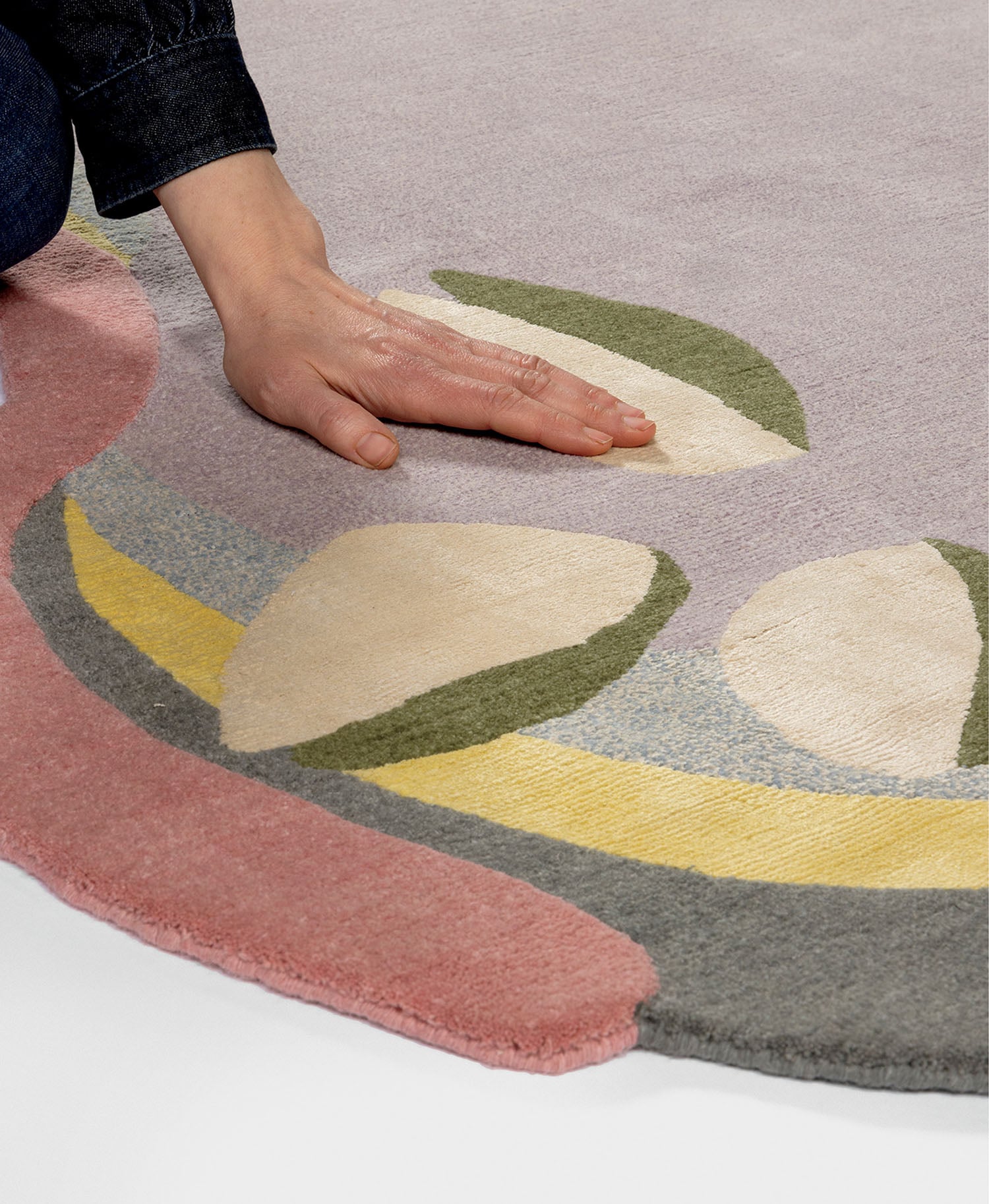 A hand touches a multicolored, patterned rug with curved shapes in pink, yellow, green, gray, and cream on a light background.