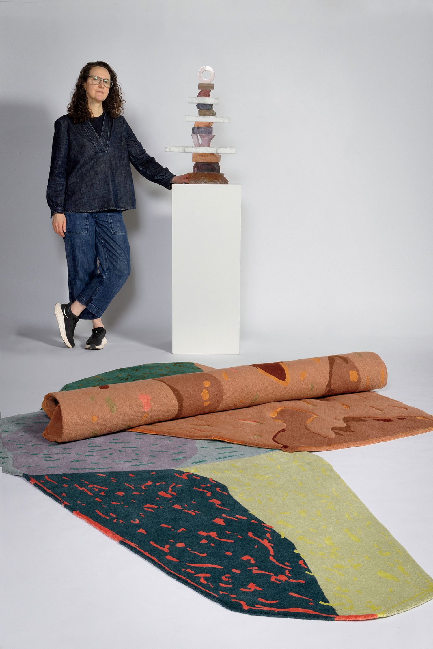 A person stands beside a white pedestal holding a stacked abstract sculpture, with colorful patterned rugs rolled and spread on the floor in front of them.