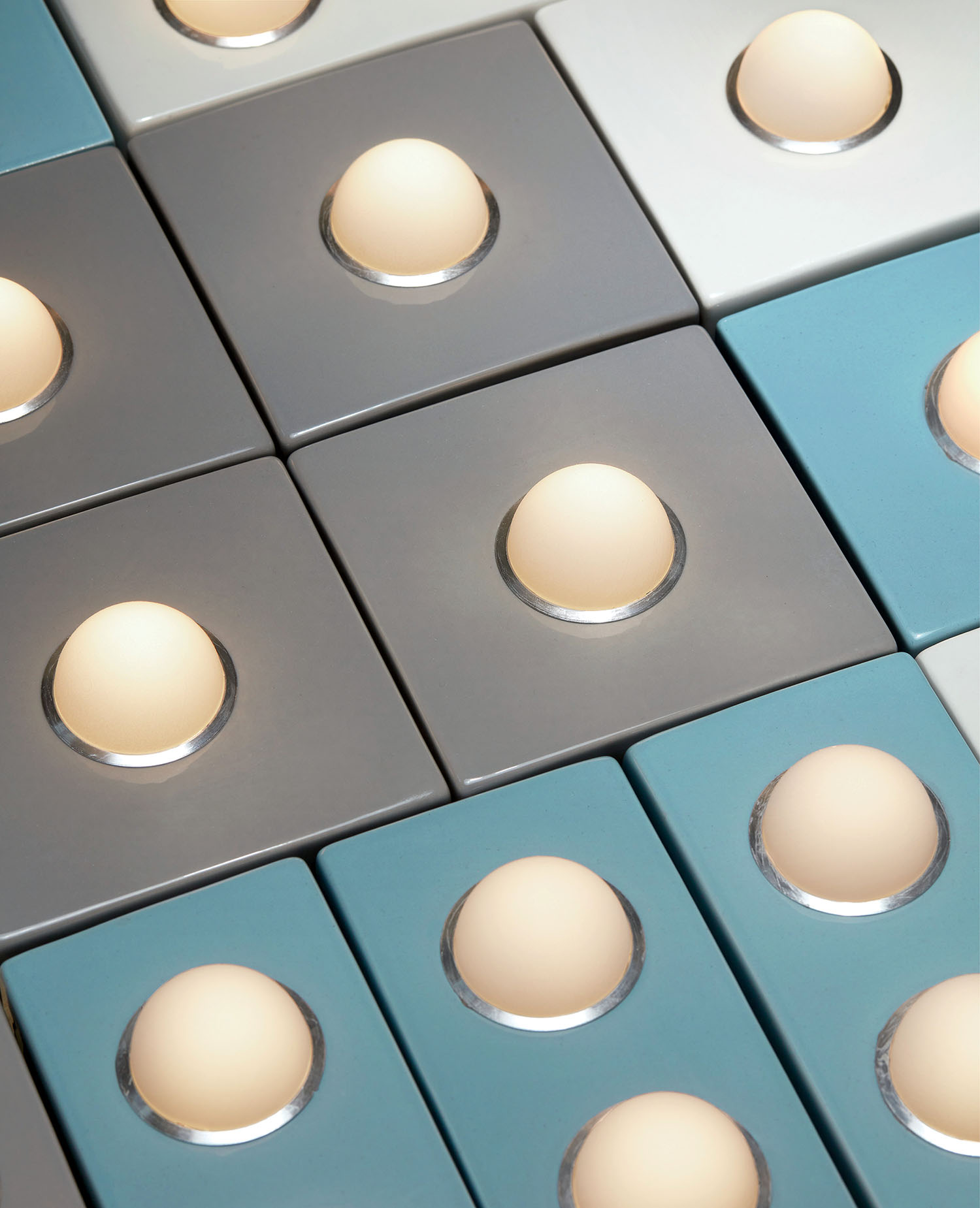 A close-up of a grid of rectangular tiles in shades of gray and blue, each featuring a single egg placed in a circular indentation at the center.