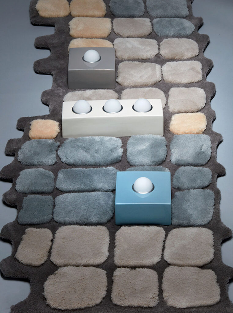 A textured rug with square patches in gray, beige, and cream tones, featuring three rectangular objects with dome-shaped buttons on top arranged in a line.