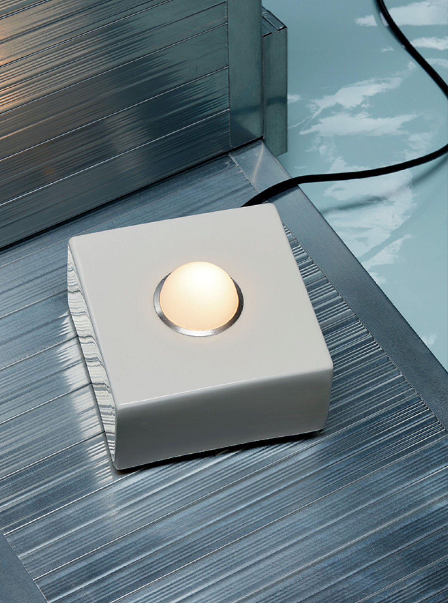 A small, square white lamp with a round bulb in the center sits on a metallic, ribbed surface with a power cord extending from its side.
