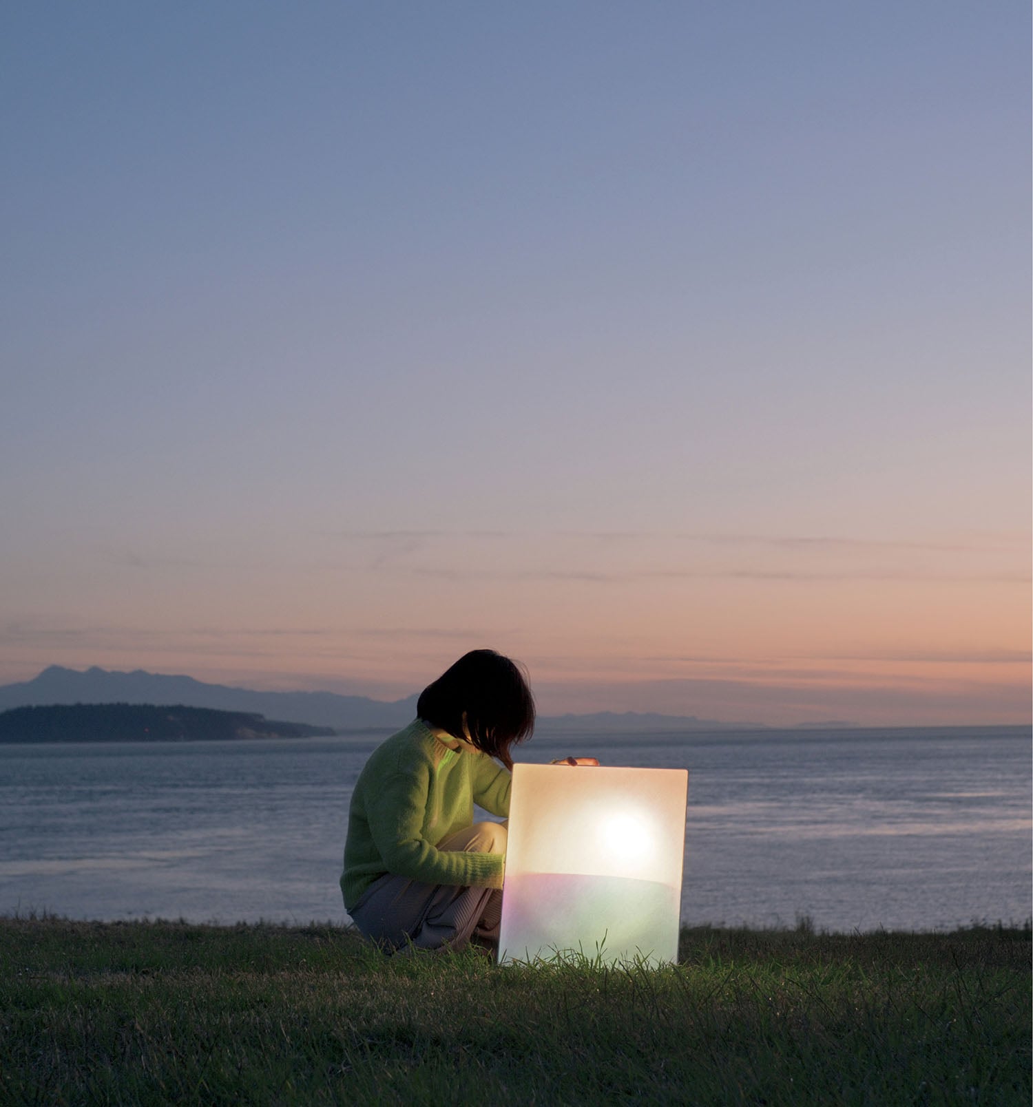 A person in a green sweater sits on grass by the water at dusk, looking at a large, illuminated lantern.