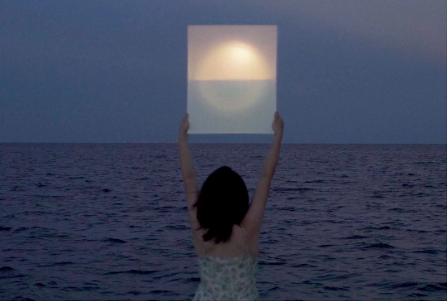 A person stands at the edge of the sea at dusk, holding a translucent square panel that frames the setting sun and horizon.