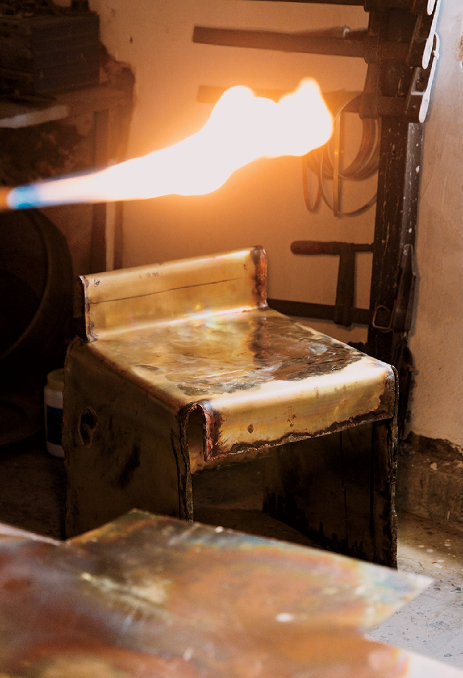 A metal chair with a worn surface is in a workshop as a bright flame from a torch passes in front of it.