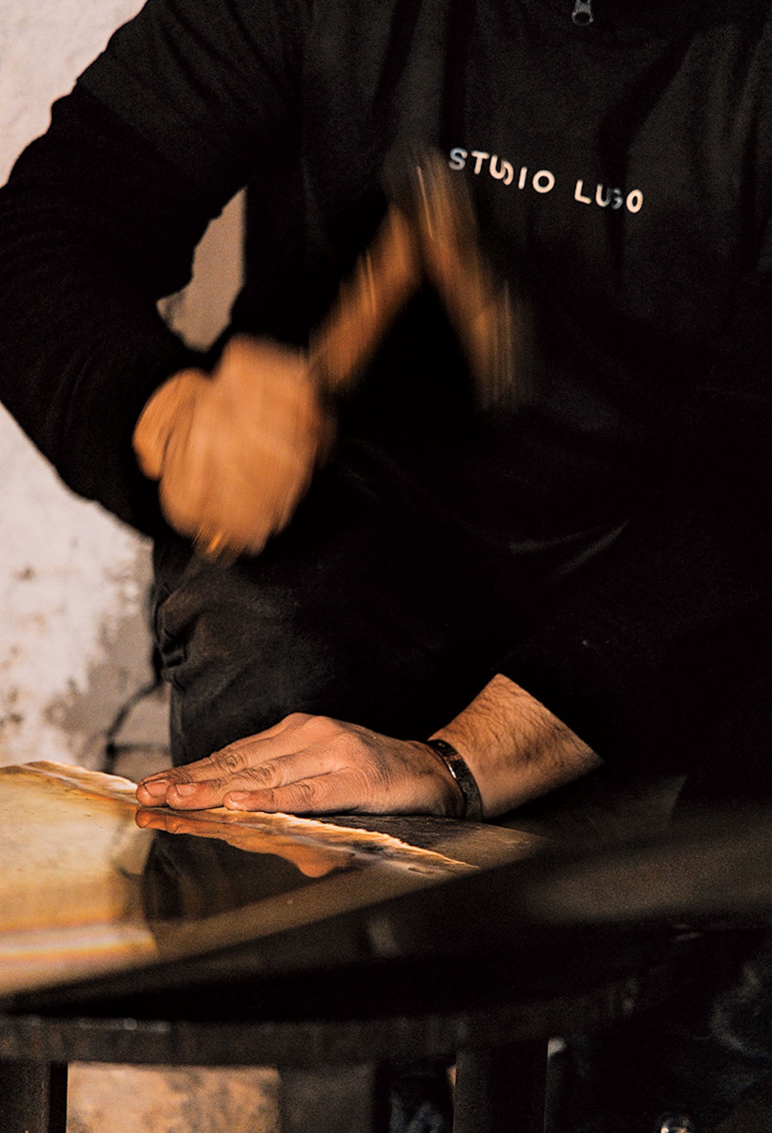 A person wearing a black "STUDIO LUDO" shirt uses a hammer to work on a flat surface, with one hand steadying the material.
