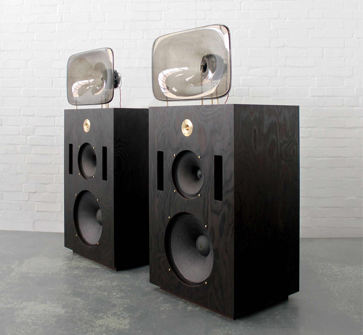Two large, dark wood speakers with prominent round drivers and unique, clear horn structures on top, set against a white brick wall.