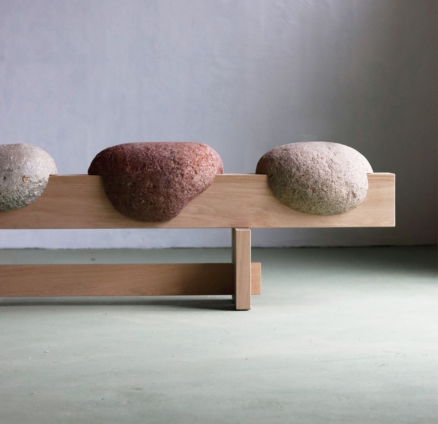 A wooden bench with three smooth, rounded stone seats in varying colors, positioned against a minimalist light gray wall and green floor.