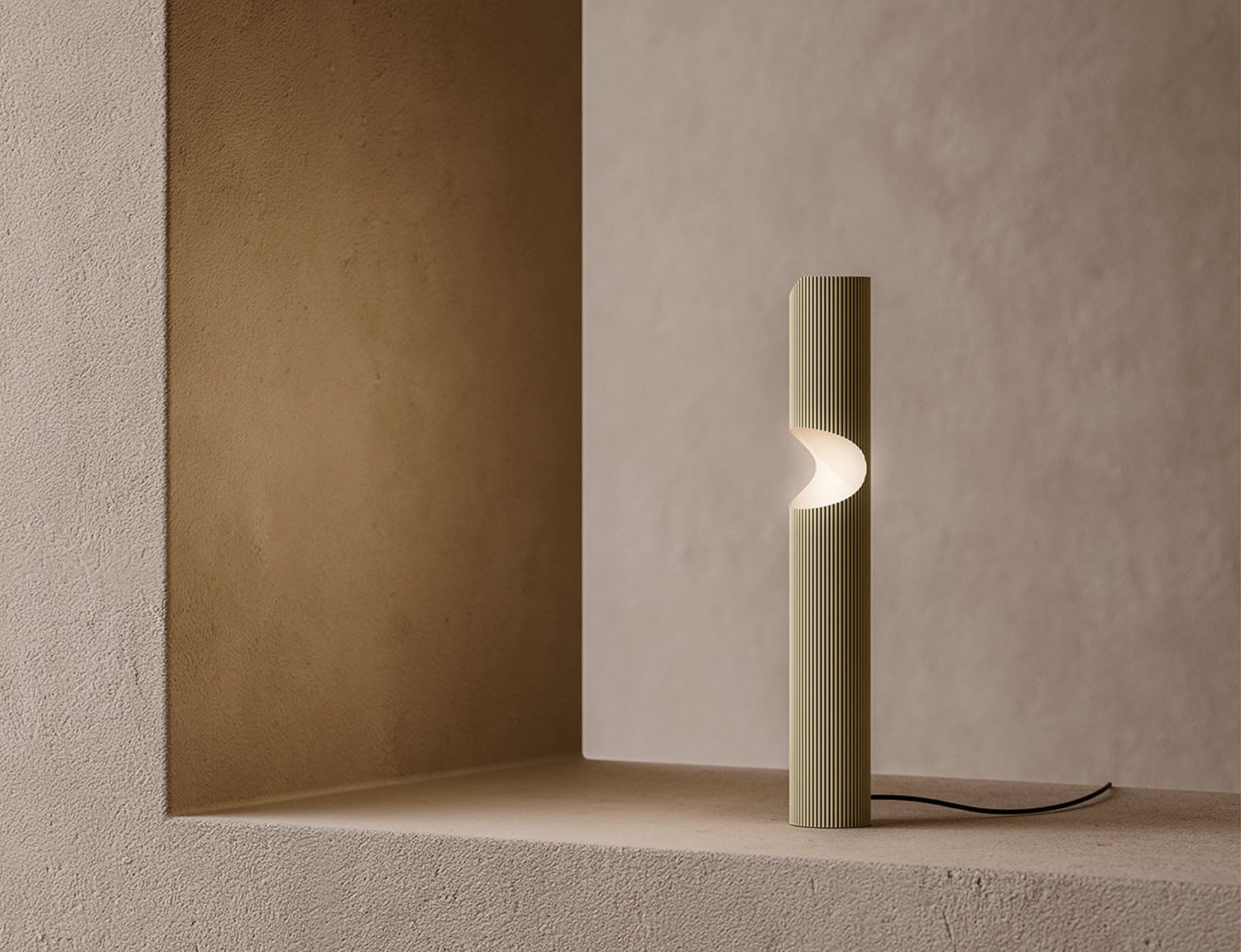 A modern cylindrical table lamp with a vertical ribbed texture emits light from a crescent-shaped cutout, placed on a beige textured surface near a wall.
